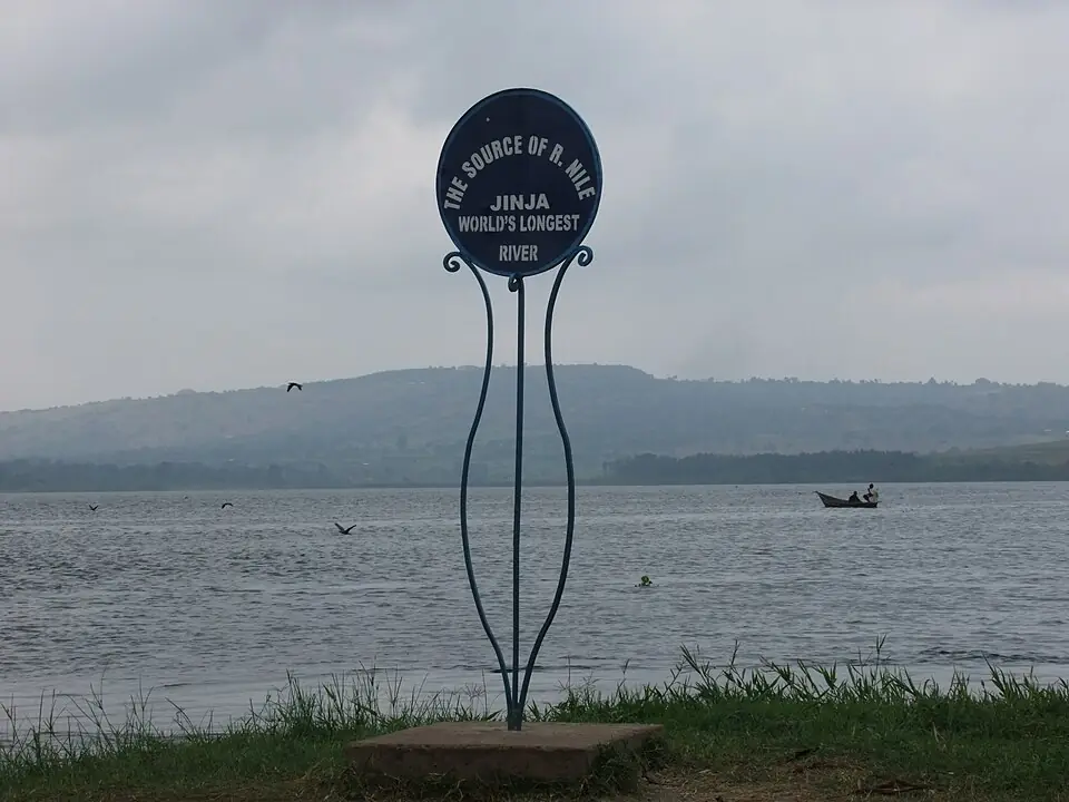 Синя табела „The Source of R. Nile – Jinja“ на брега, зад нея широката вода и рибарска лодка